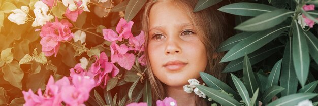 Portrait face of candid beautiful little kid girl of eight years old on background of green plants and pink flowers during a summer vacation travel gen z mental health concept banner flare