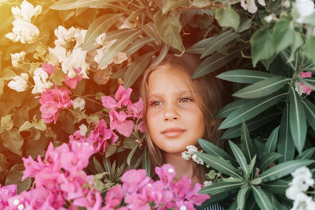 Portrait face of candid beautiful little kid girl of eight years old with brown eyes on background of green plants and pink flowers during a summer vacation travel gen z mental health concept flare