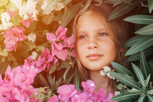 Portrait face of candid beautiful little kid girl of eight years old with brown eyes on background of green plants and pink flowers during a summer vacation travel gen z mental health concept flare