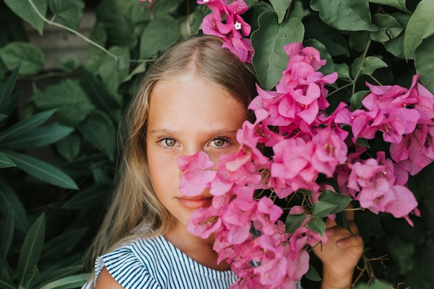 Portrait face of candid beautiful little kid girl of eight years old with green brown eyes on background of green plants and pink flowers during a summer vacation travel gen z mental health concept