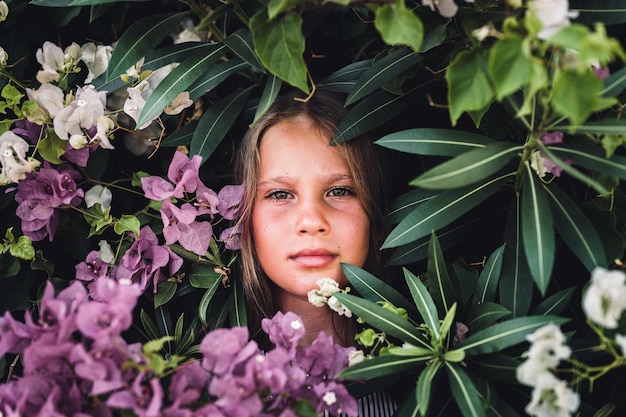 Portrait face of candid beautiful little kid girl of eight years old with green brown eyes on background of green plants and pink flowers during a summer vacation travel gen z mental health concept