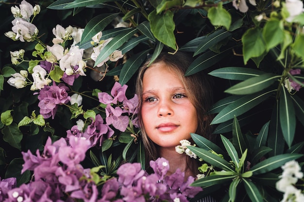 Portrait face of candid beautiful little kid girl of eight years old with green brown eyes on background of green plants and pink flowers during a summer vacation travel gen z mental health concept