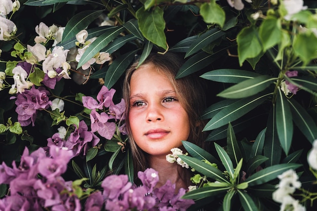 Portrait face of candid beautiful little kid girl of eight years old with green brown eyes on background of green plants and pink flowers during a summer vacation travel gen z mental health concept