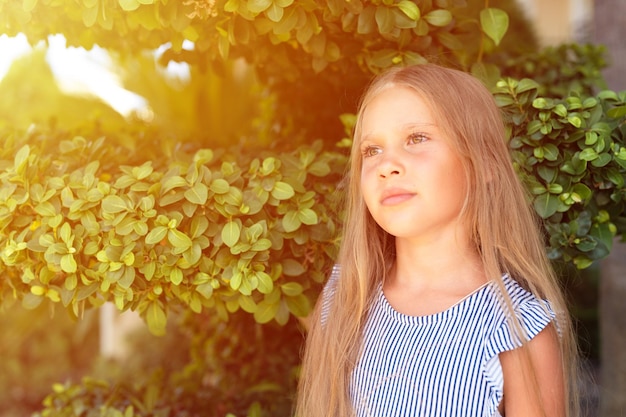 Portrait face of candid happy little kid girl of eight years old with long blond hair and green eyes on background of green plants during a summer vacation travel gen z mental health concept flare