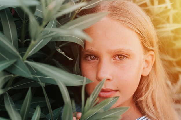 Portrait face of candid happy little kid girl of eight years old with long blond hair and green eyes on background of green plants during a summer vacation travel gen z mental health concept flare