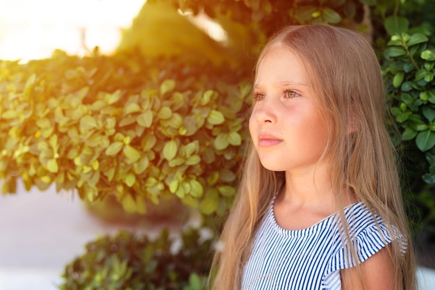 Portrait face of candid happy little kid girl of eight years old with long blond hair and green eyes on background of green plants during a summer vacation travel gen z mental health concept flare