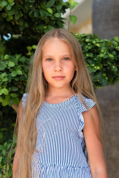 Portrait face of candid happy little kid girl of eight years old with long blond hair and green eyes on background of green plants during a summer vacation travel gen z mental health concept