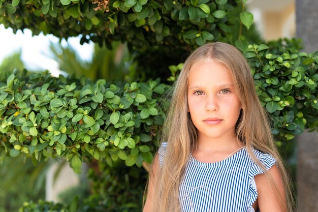 Portrait face of candid happy little kid girl of eight years old with long blond hair and green eyes on background of green plants during a summer vacation travel gen z mental health concept