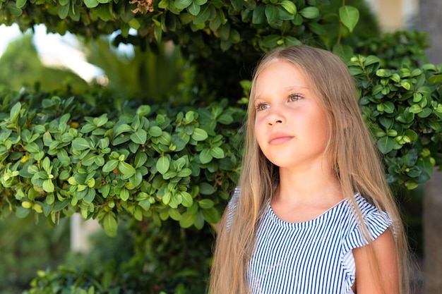 Portrait face of candid happy little kid girl of eight years old with long blond hair and green eyes on background of green plants during a summer vacation travel gen z mental health concept