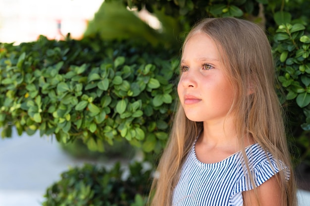 Portrait face of candid happy little kid girl of eight years old with long blond hair and green eyes on background of green plants during a summer vacation travel gen z mental health concept