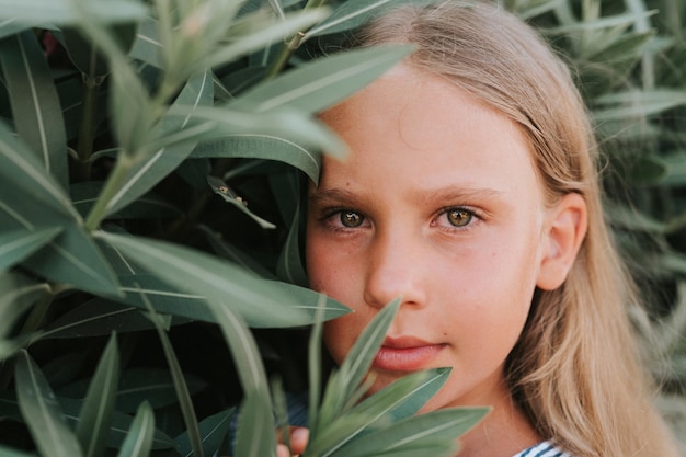 Portrait face of candid happy little kid girl of eight years old with long blond hair and green eyes on background of green plants during a summer vacation travel gen z mental health concept