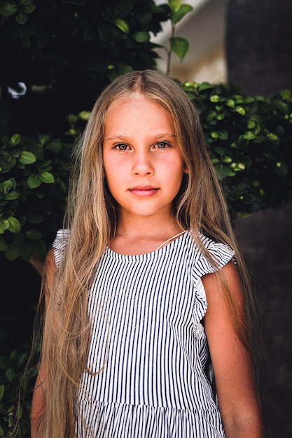 Portrait face of candid happy little kid girl of eight years old with long blond hair and green eyes on background of green plants during a summer vacation travel gen z mental health concept