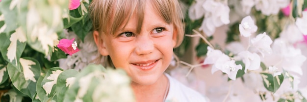 Portrait of face candid little happy smiling five year old blonde kid boy with green eyes in pink and white flower plants in nature children have fun summer holidays bright light and airy banner