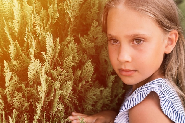 Portrait face of candid offended upset little kid girl of eight years old with long blond hair on background of green plants during a summer vacation travel gen z mental health concept flare