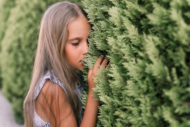 Portrait face of candid offended upset little kid girl of eight years old with long blond hair and green eyes on background of green plants during a summer vacation travel gen z mental health concept