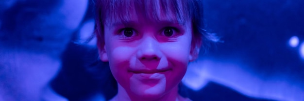 Portrait face cute happy candid caucasian little six year old kid boy with long blonde hair in the studio illuminated by blue purple neon light banner