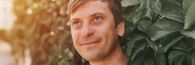 Portrait of face happy smiling unshaven mature 40 years man solo traveler with stubble on the background of green trees plants in nature on sunny summer day on vacation travel banner flare