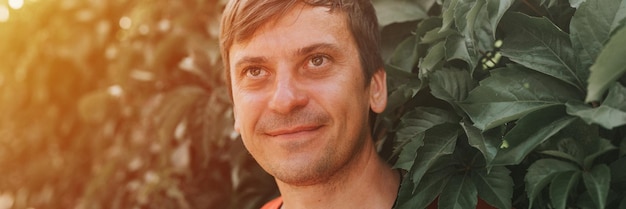 Portrait of face happy smiling unshaven mature 40 years man solo traveler with stubble on the background of green trees plants in nature on sunny summer day on vacation travel banner flare