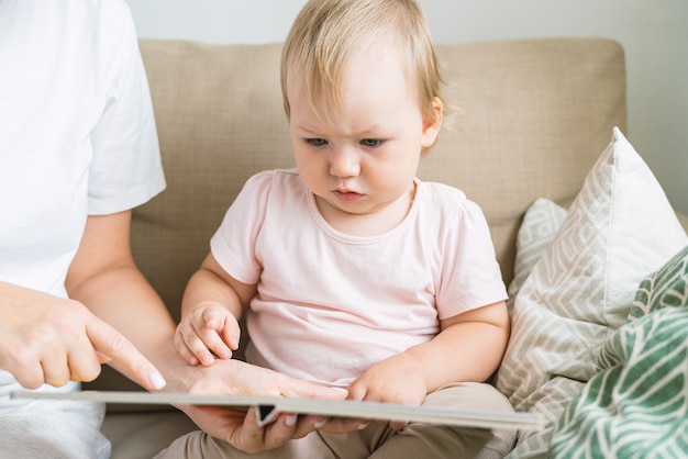 Portrait of faceless female pointing out in book to serious blond child sitting on sofa close up