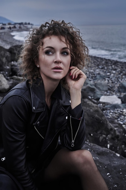 Portrait of fashion model girl in black jacket sitting on a stone by the sea