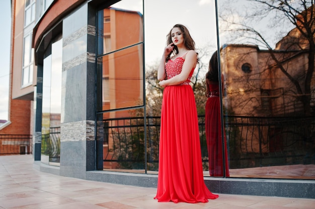 Portrait of fashionable girl at red evening dress posed background mirror window of modern building