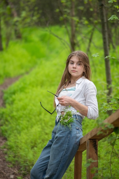 Portrait of a fashionable little girl standing leaning against a wooden village bridge against the background of green trees of the forest