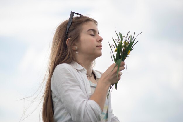 Portrait of a fashionable little girl with a bouquet of wildflowers in a green field against a background of a cloudy sky