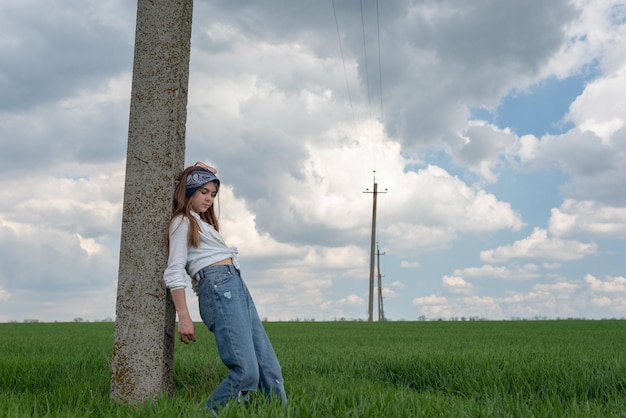 Portrait of a fashionable little teenage girl posing by a pillar in a green grass field rural landscape in jeans