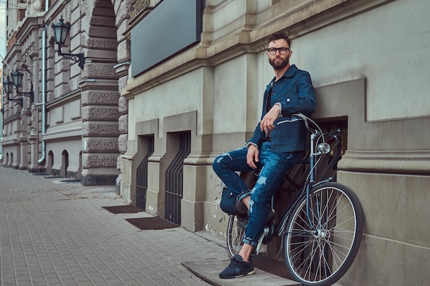 Portrait of a fashionable man in stylish clothes leaning against a wall with city bicycle on the street.