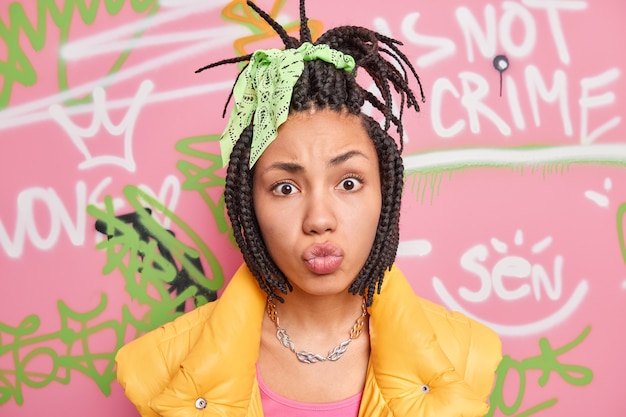 Portrait of fashionable teenage girl make grimace at camera pouts lips has combed dreadlocks belongs to youth subculture poses in urban place against colorful drawn graffiti wall