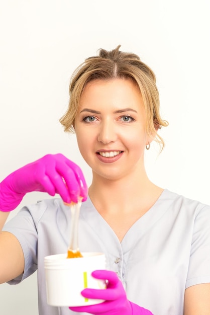 Portrait of a female caucasian beautician holding a jar of sugar paste for sugaring wearing pink gloves on white background