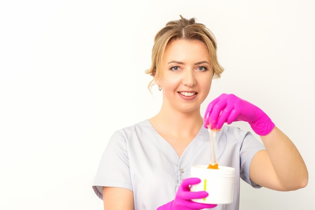 Portrait of a female caucasian beautician holding a jar of sugar paste for sugaring wearing pink gloves on white background