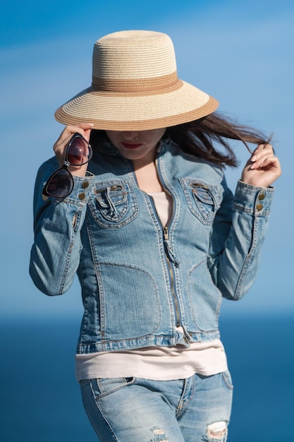 Portrait of female in denim jacket and jeans hiding with straw hat over his face