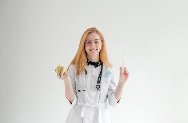 Portrait of a female doctor standing with an apple and a syringe on a light background