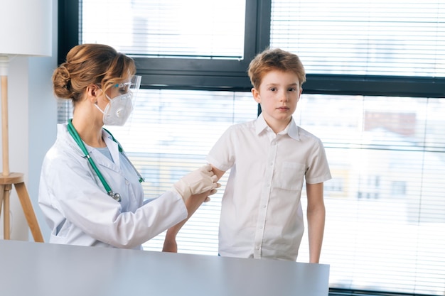 Portrait of female doctor wearing white uniform applying plaster on shoulder of child boy after vaccination injection by window