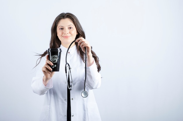 Portrait of female doctor with stethoscope and cup of coffee standing on white wall.