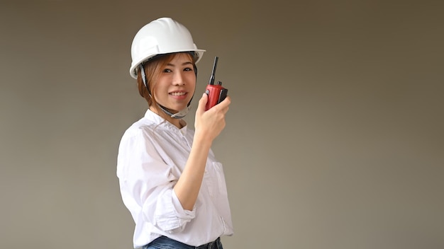 Portrait of female engineer in a protective helmet standing against grey background and looking at camera