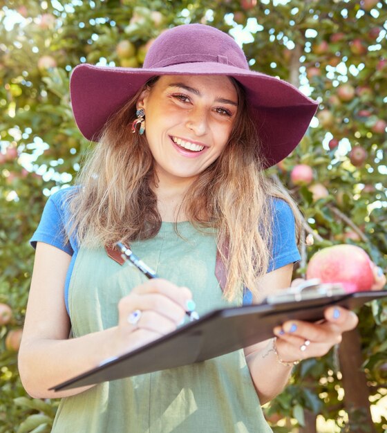 Portrait of female farm worker holding an apple while writing and making notes on a orchard farm during harvest season Agronomist doing inspection and record keeping on the quality of produce