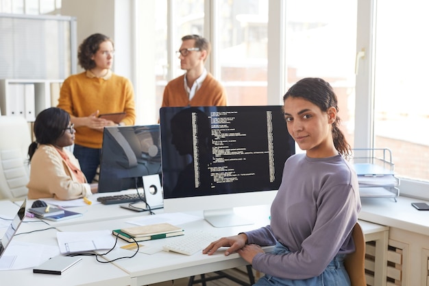 Portrait of female IT developer looking at camera while posing against computer with code on screen in software production studio, copy space