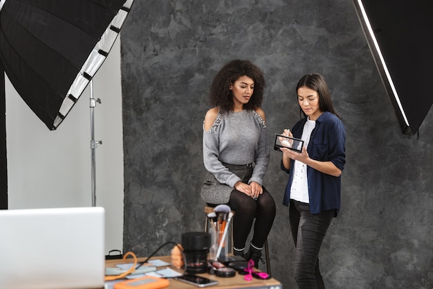 Portrait of female makeup artist applying cosmetics to charming woman during photo shooting with professional camera and softbox in studio