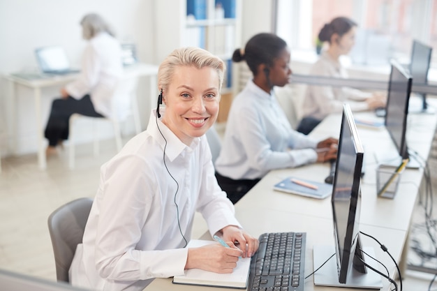 Portrait of female operator wearing headset and smiling while sitting in row in call center or office interior