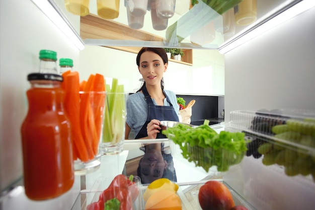 Portrait of female standing near open fridge full of healthy food vegetables and fruits