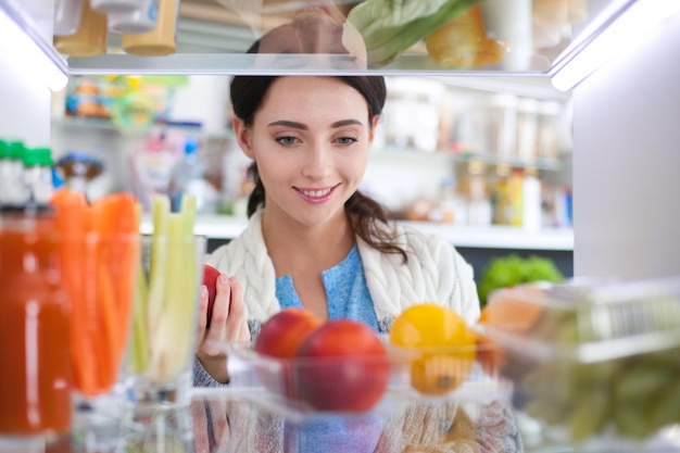 Portrait of female standing near open fridge full of healthy food vegetables and fruits