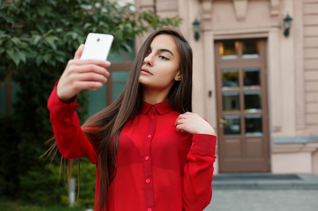 Portrait female in stylish outfit making selfie. Portrait of beautiful young woman visiting a destination city , filming herself in selfie video using mobile phone, waving, smiling looking at camera.