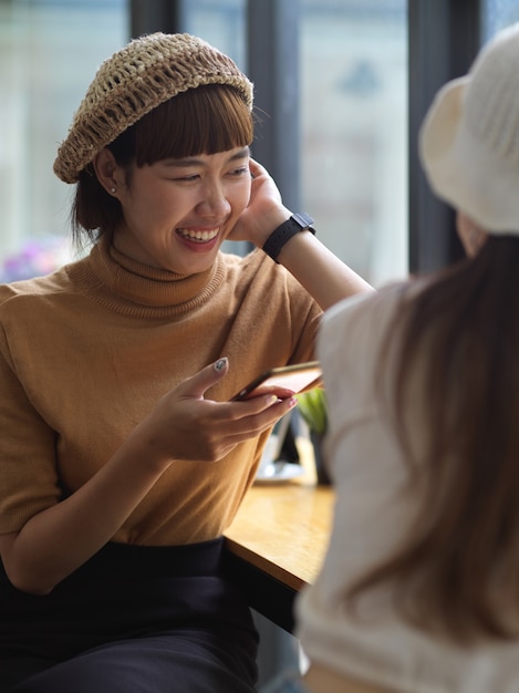 Portrait of female teenager using smartphone and talking with friend while sitting in cafe