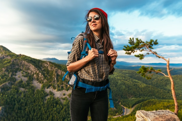 Portrait of a female traveler in the mountains. Adventure, travel and hiking concept. A happy woman in a red cap enjoying the sunlight during a hike in the mountains. Female tourist with glasses