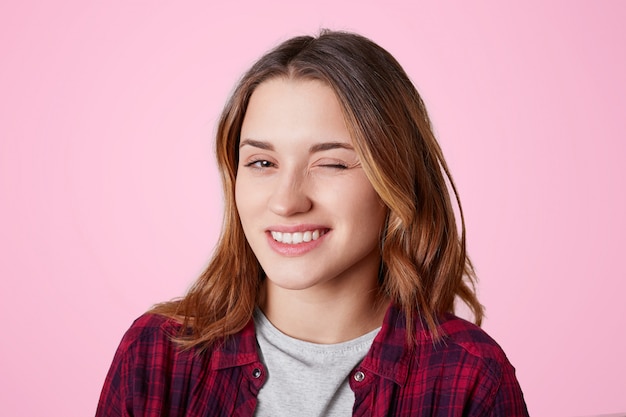 Portrait of female with cheerful expression, blinks eye as flirts with handsome man, expresses her sympathy, dressed in casual checkred shirt, isolated on pink. Woman winks indoor