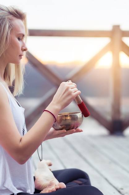 Portrait of Female yoga teacher playing tibetan bowl or singing bell on a wooden bridge