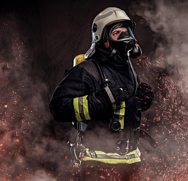 Portrait of firefighter dressed in uniform and an oxygen mask standing in fire sparks and smoke over a dark background.