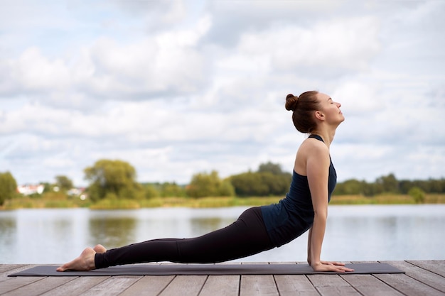 Portrait of fit sporty young woman practicing yoga on yoga mat on the wooden pier near lake.